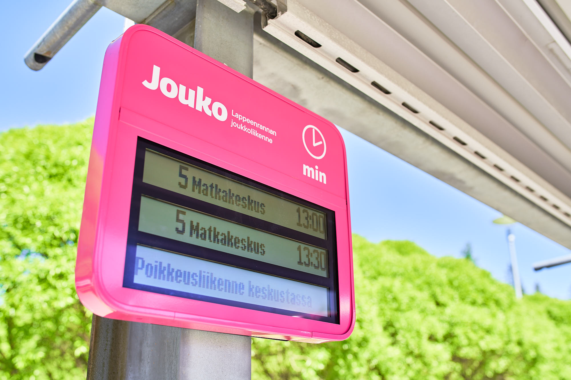 Pinkki Jouko-joukkoliikenteen sähköinen opastekyltti bussipysäkillä, ruudussa teksti ”5 Matkakeskus 13:00 ja 13:30, poikkeusliikenne keskustassa”.